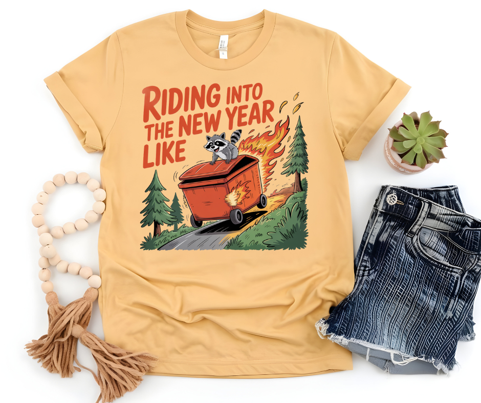 Graphic T-Shirt With A Raccoon Riding On A Dumpster Fire Down A Hill With The Text Riding Into The New Year Like